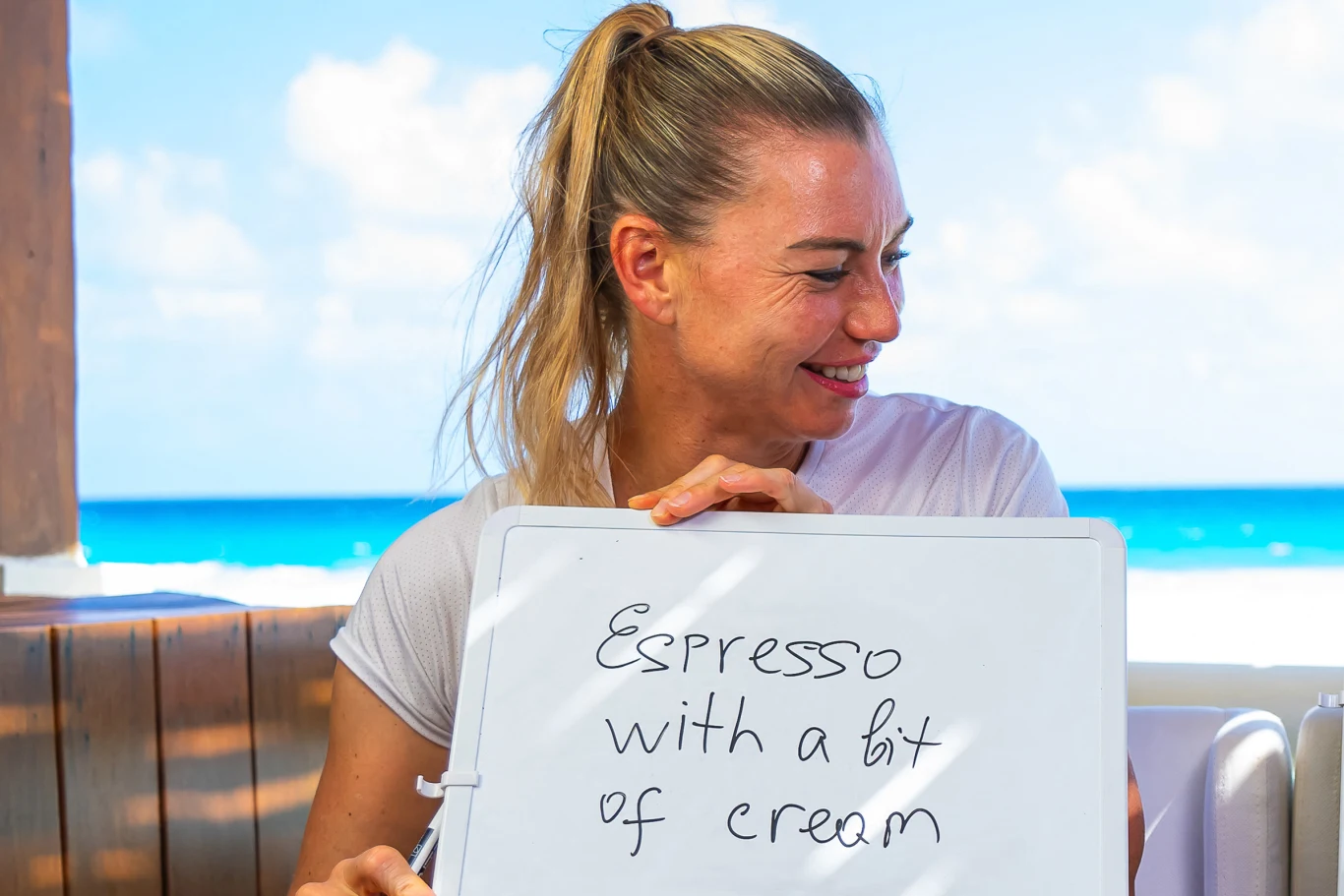 Wiera Zwonariowa uśmiechnięta kobieta z blond włosami spiętymi w kucyk siedzi nad morzem i trzyma tabliczkę z napisem „Espresso with a bit of cream”
