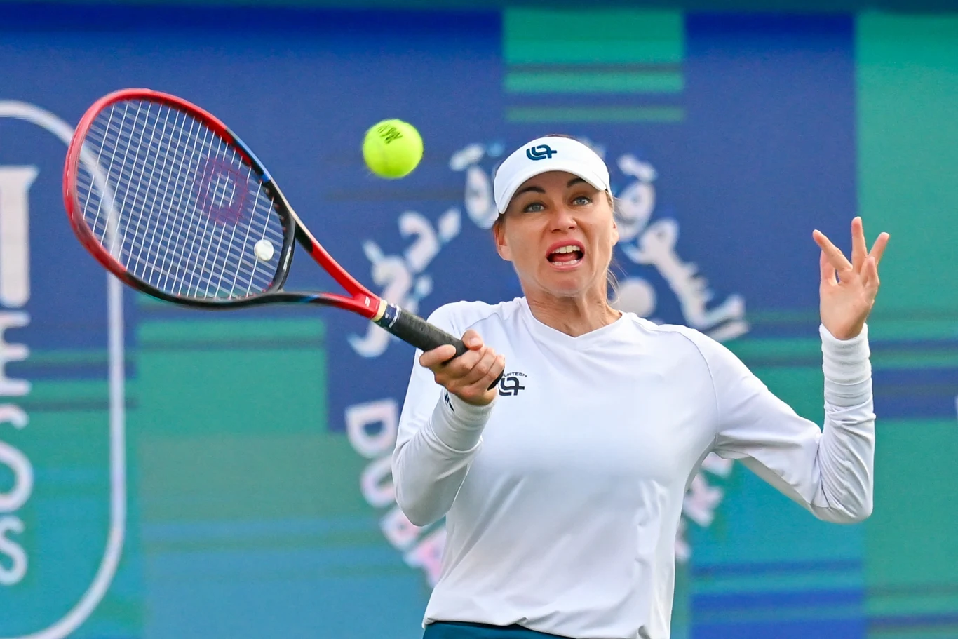 tenisistka w białej koszulce i czapce uderza żółtą piłkę tenisową rakietą podczas meczu na tle niebiesko-zielonego baneru z białym napisem i grafiką