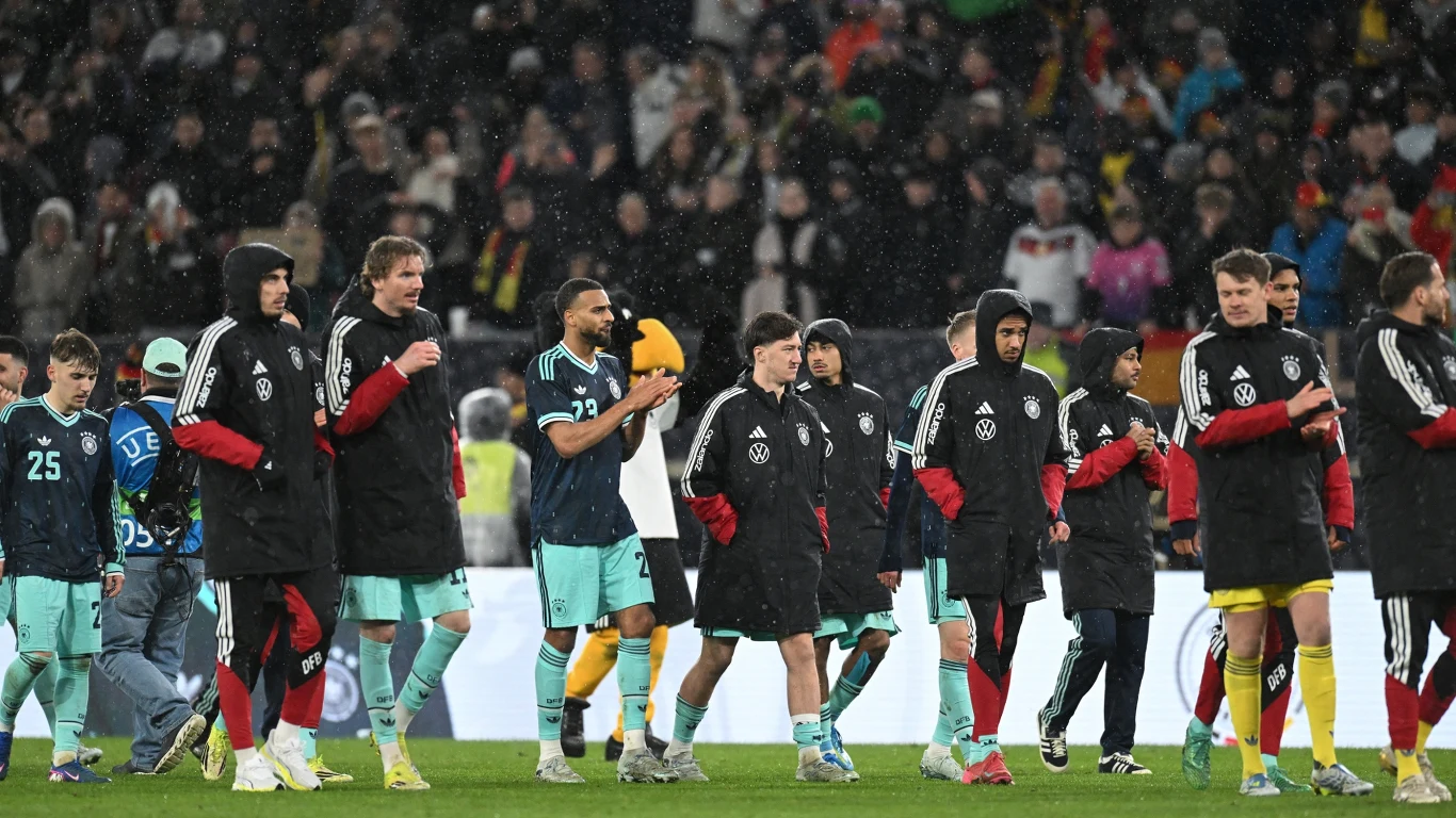 piłkarze w sportowych strojach oraz czarnych kurtkach maszerują po murawie stadionu, wokół nich znajdują się inni członkowie zespołu i sztabu, w tle widoczni są kibice na trybunach