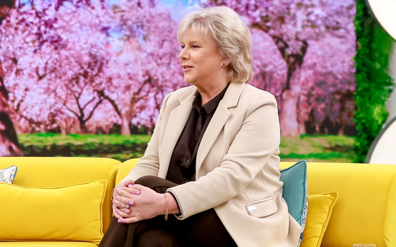 Dorota Kamińska w "halo tu polsat" Kobieta o jasnych włosach w eleganckim, kremowym żakiecie siedzi na żółtej sofie, w tle kolorowe wiosenne drzewa pełne różowych kwiatów.