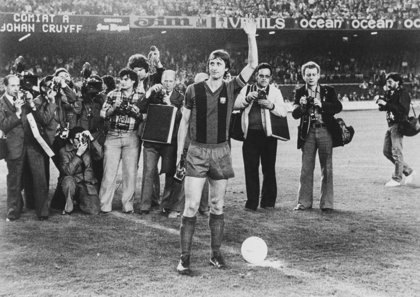 Piłkarz w stroju FC Barcelony stoi na środku boiska z uniesioną ręką, wokół niego grupa fotografów robiących zdjęcia, w tle widoczna tablica świetlna z napisem oraz tłum na trybunach.