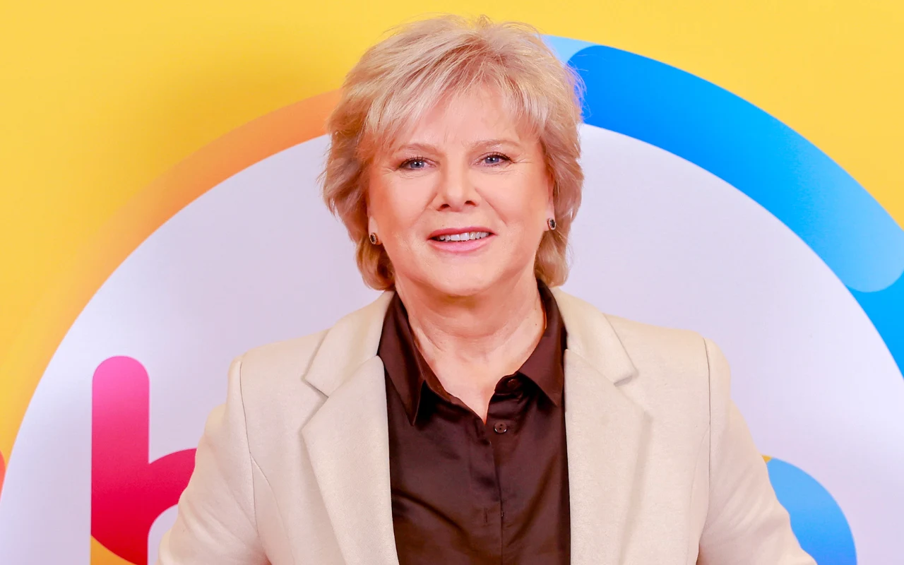 Dorota Kamińska świętuje 50 lat od swojego ekranowego debiutu Uśmiechnięta kobieta o jasnych włosach w eleganckiej, beżowej marynarce i ciemnej koszuli na tle kolorowej, graficznej ściany z żółtym, pomarańczowym i niebieskim akcentem.