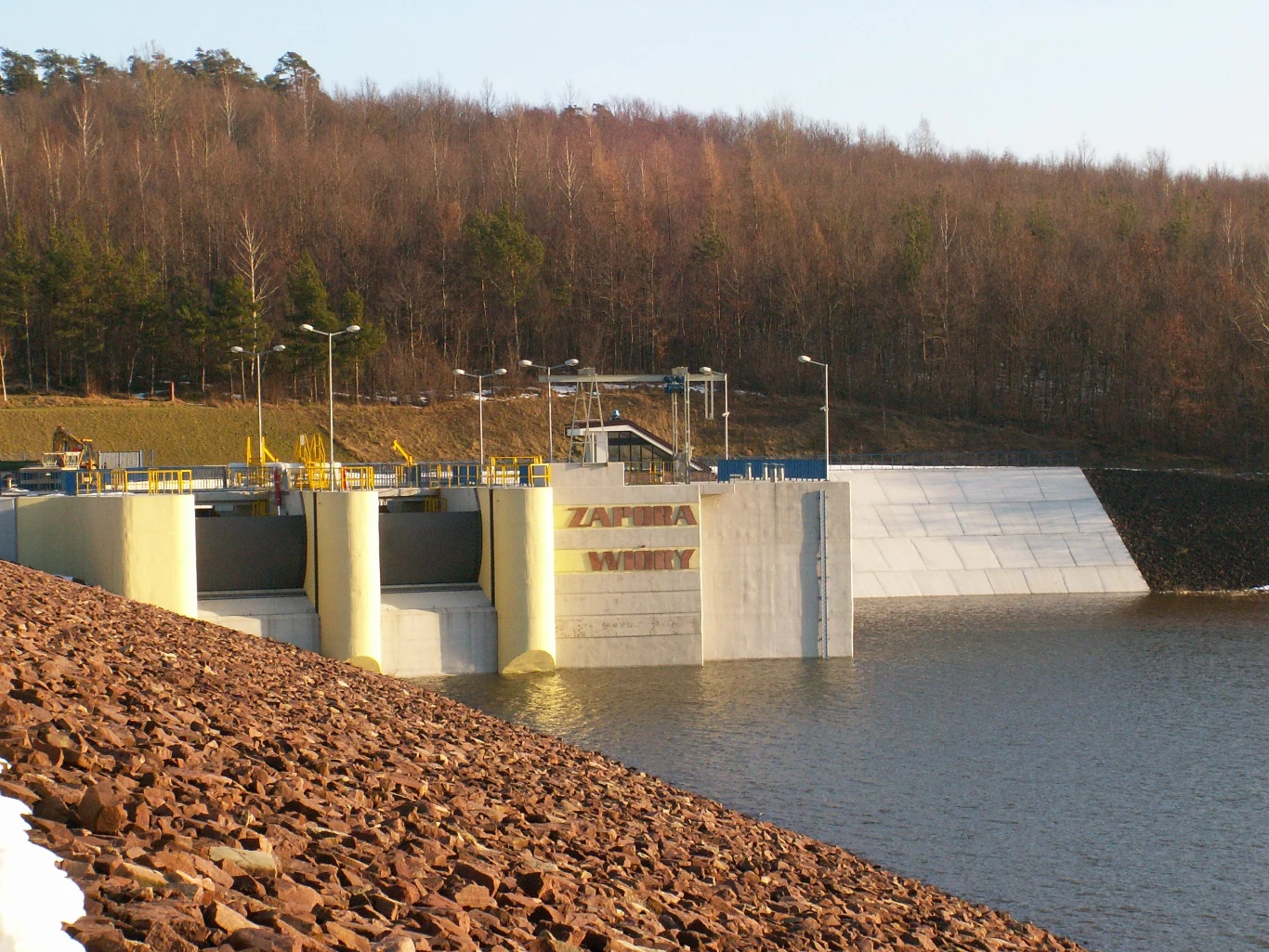 betonowa zapora wodna z wyraźnym napisem „Zapora Wióry”, otoczona zbiornikiem wodnym, w tle porośnięte drzewami wzgórze, metalowe barierki oraz elementy infrastruktury hydrotechnicznej na koronie zapory