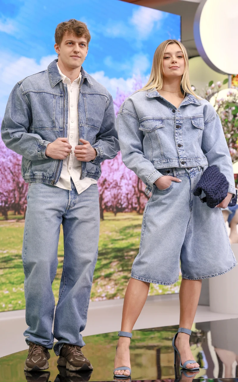 dwoje młodych ludzi ubranych w modne, jasnoniebieskie ubrania z dżinsu – mężczyzna w kurtce jeansowej, jasnej koszuli i prostych jeansach, kobieta w obszernej, krótkiej kurtce jeansowej, szerokich spodenkach z dżinsu i sandałach na obcasie, stoją na tl...