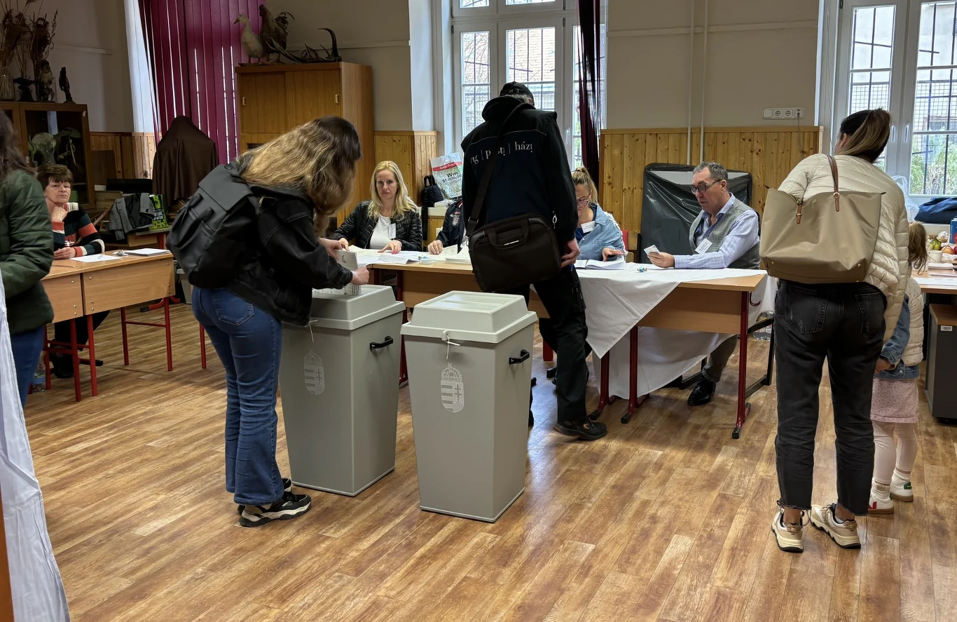 Ludzie oddający głosy wrzucając karty do urn wyborczych, komisja siedząca przy stole w tle, wnętrze sali z drewnianą podłogą i dużymi oknami.