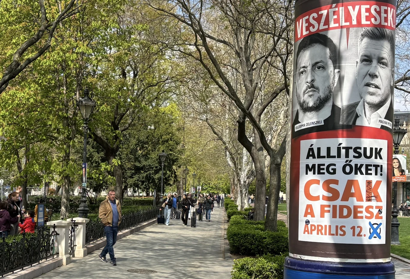 Duży plakat wyborczy z dwoma mężczyznami oraz wyrazistym napisem w języku węgierskim przytwierdzony do latarni na tle alejki miejskiej z przechodniami, otoczonej drzewami w parku.