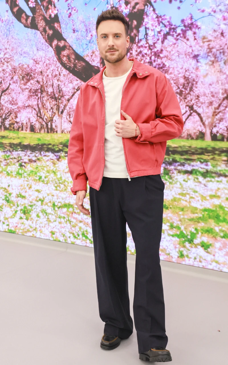 mężczyzna w jasnej koszulce i czerwonej kurtce stoi na tle kwitnących drzew, ubrany w ciemne, szerokie spodnie i czarne buty, z wyprostowaną sylwetką i lekko uniesioną ręką