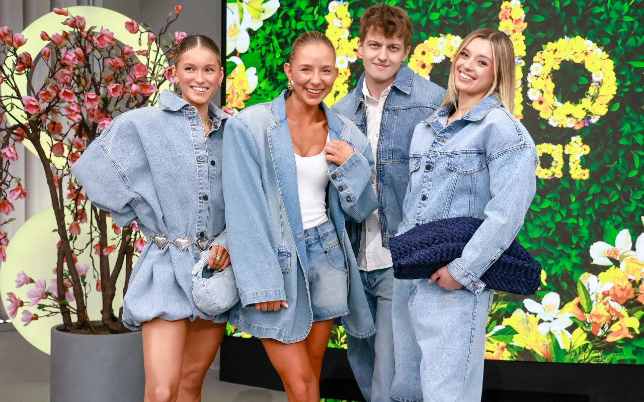 cztery młode osoby ubrane w luźne, jasnoniebieskie stroje z dżinsu stoją razem i uśmiechają się do kamery, w tle kwitnące gałęzie i kolorowa dekoracja z kwiatami na zielonej ścianie