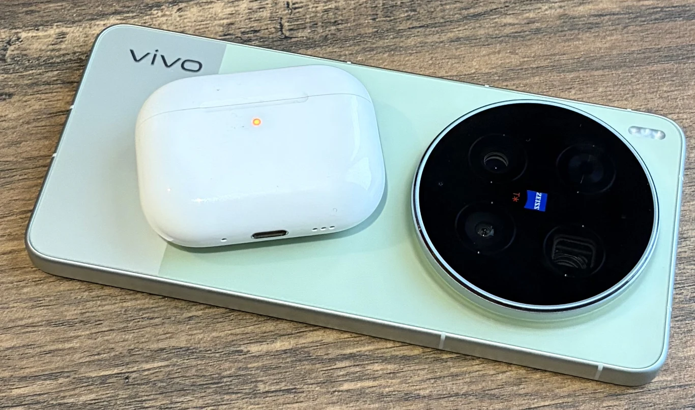 smartfon vivo z dużym modułem aparatów z obiektywem sygnowanym logo Zeiss, obok niego leżące na obudowie białe bezprzewodowe etui na słuchawki na drewnianym blacie