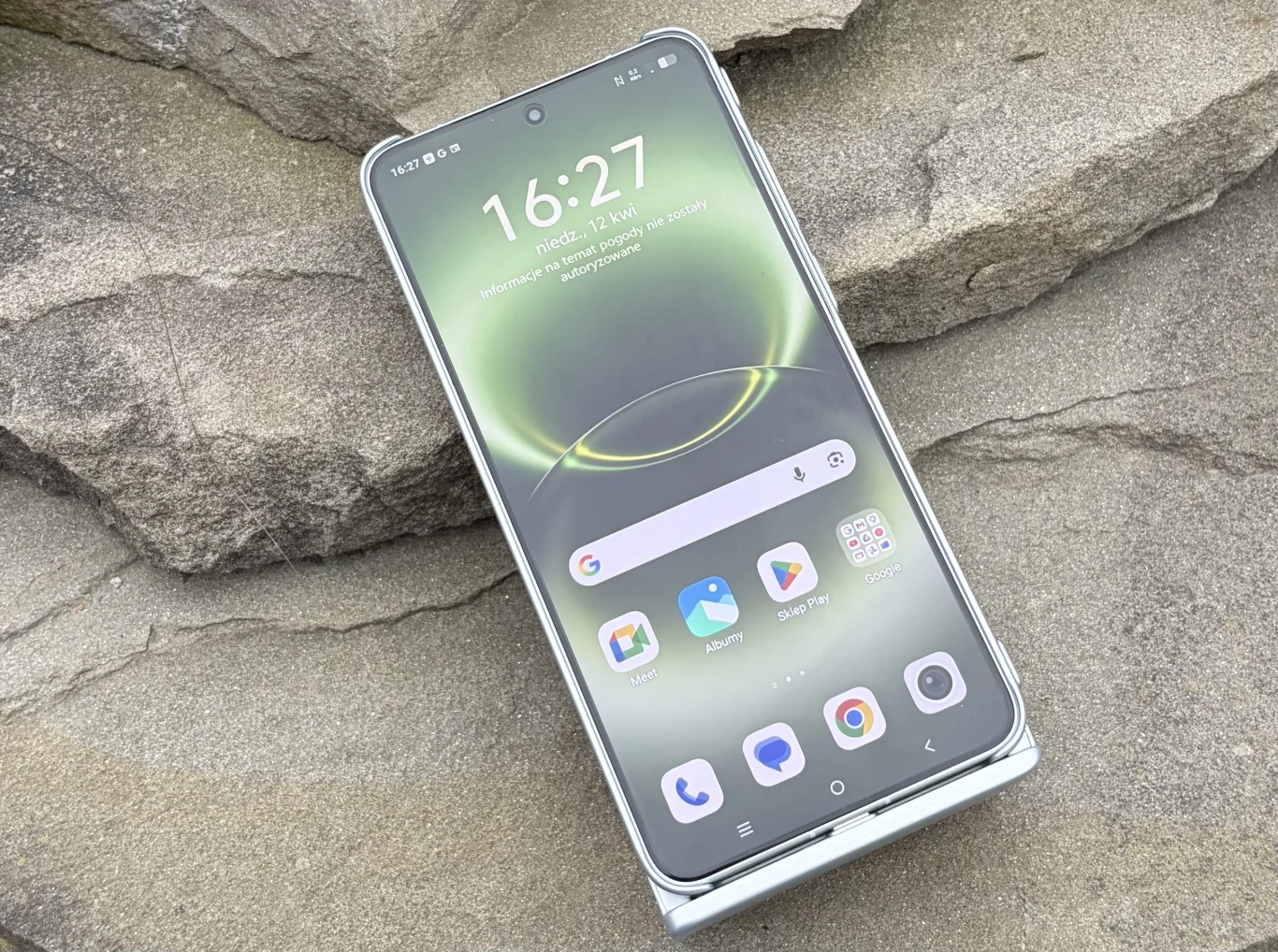 nowoczesny smartfon z dużym wyświetlaczem AMOLED, wyraźnie widocznymi ikonami aplikacji i cienkimi ramkami, położony na jasnoszarej, kamiennej powierzchni