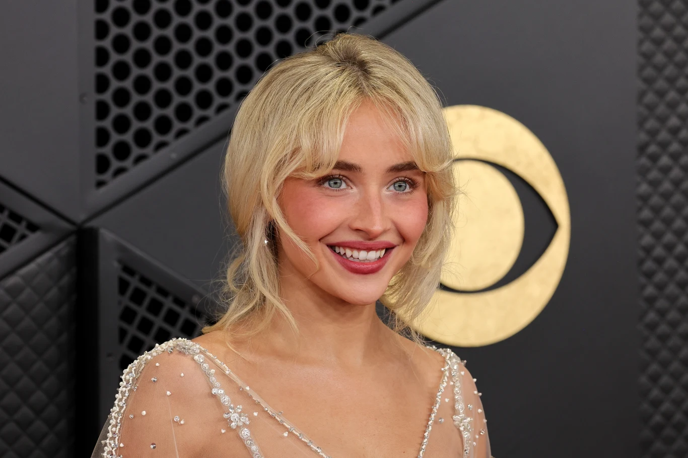 uśmiechnięta kobieta z blond włosami i niebieskimi oczami w eleganckiej, przezroczystej sukni zdobionej koralikami, pozująca na tle czarnej ścianki z geometrycznym wzorem i dużym, złotym logiem CBS