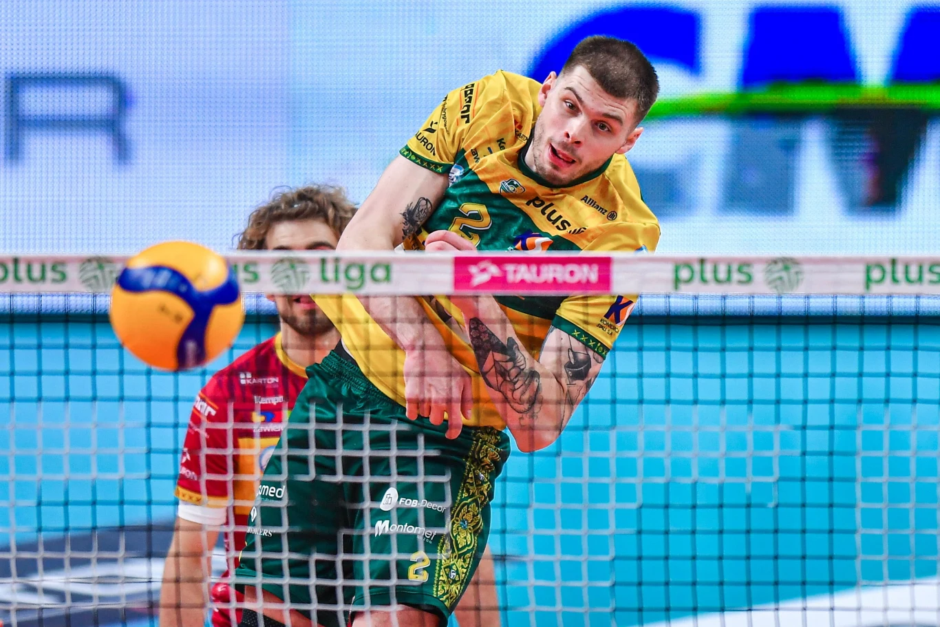 Bartosz Kwolek, przyjmujący Aluron CMC Warty Zawiercie siatkarz w żółto-zielonym stroju wykonuje dynamiczny atak nad siatką, w tle widać fragment rywala oraz rozmyty napis sponsorów