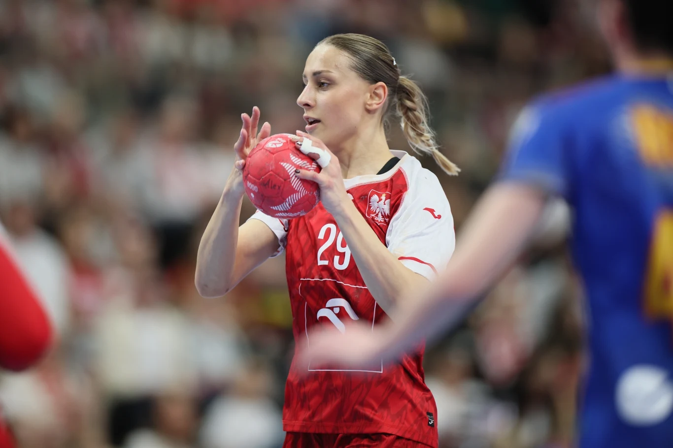 Katarzyna Cygan zawodniczka w czerwono-białym stroju z numerem 29 przygotowująca się do rzutu piłką ręczną podczas meczu, w tle rozmyta sylwetka rywalki w niebieskim stroju