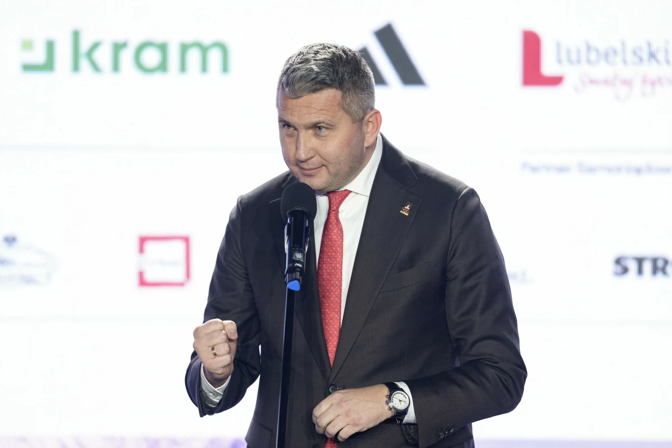 mężczyzna w garniturze i czerwonym krawacie stoi przy mikrofonie, wykonując gest zaciśniętej pięści, w tle widoczne rozmyte logotypy firm