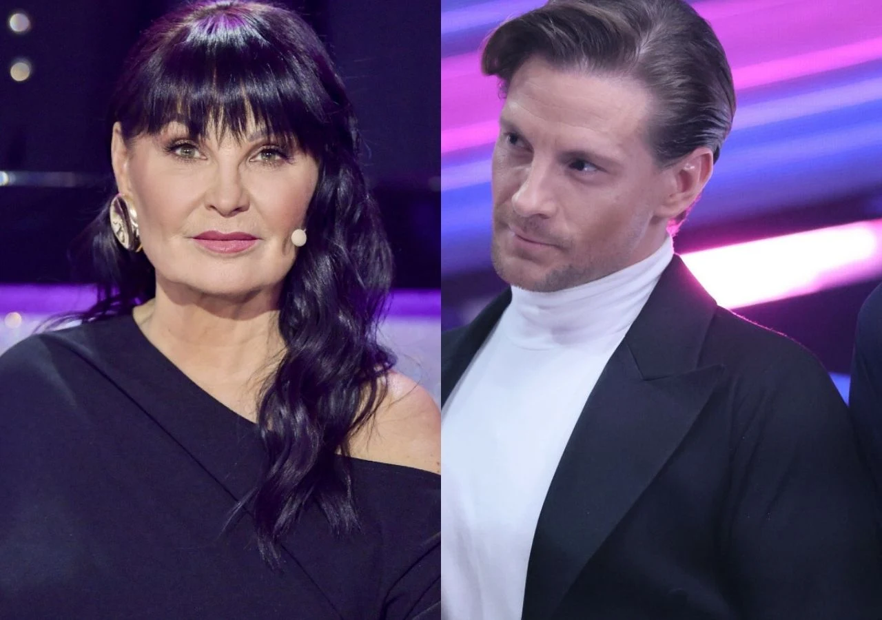 Iwona Pavlović i Sebastian Fabijański w "Tańcu z gwiazdami" Kobieta z długimi ciemnymi włosami i dużymi kolczykami w czarnej sukience patrzy prosto przed siebie, obok mężczyzna w białej koszuli i czarnym żakiecie spoglądający na nią poważnie, na tle kolorowych świateł scenicznych.