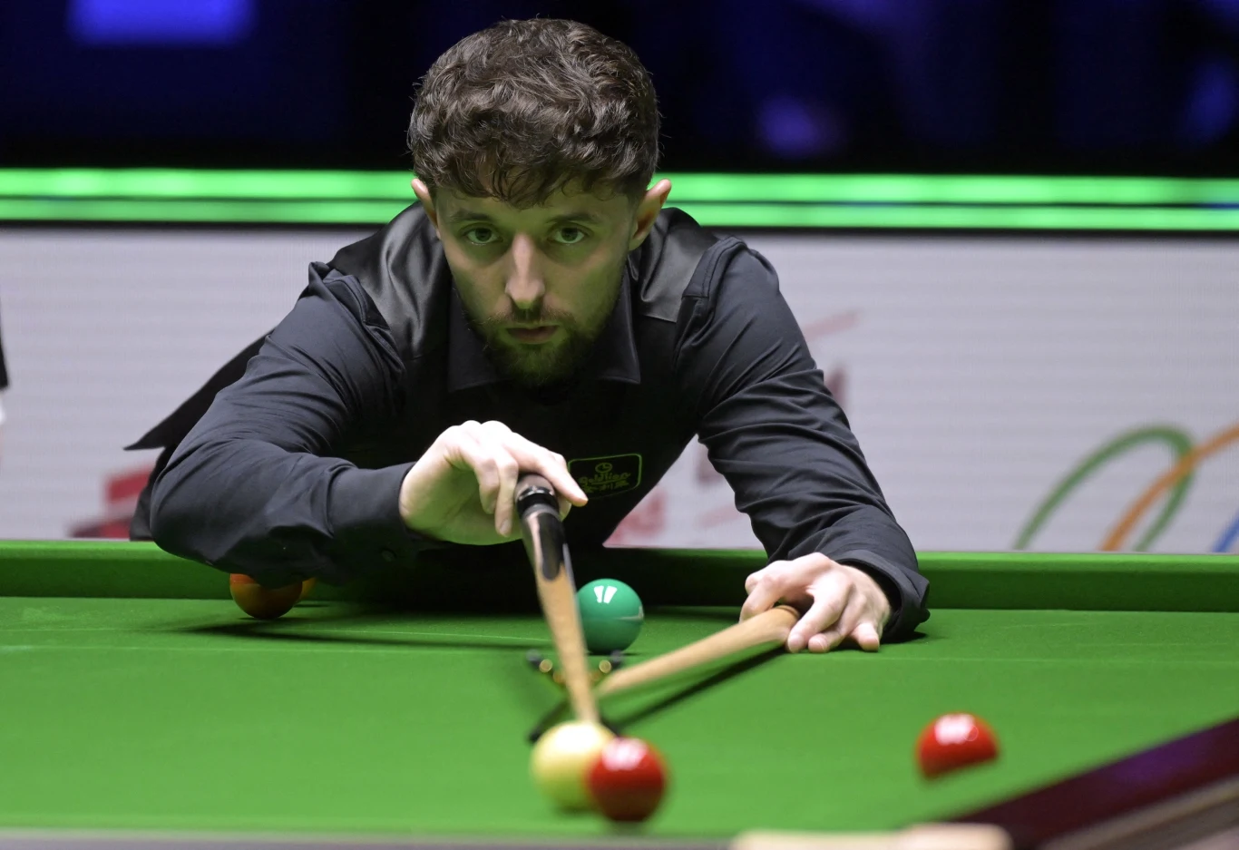 Mężczyzna w ciemnej koszuli skoncentrowany na oddaniu uderzenia kijem bilardowym, przygotowuje się do zagrania czerwonej bili na stole snookerowym; w tle rozmyte światła turniejowe.
