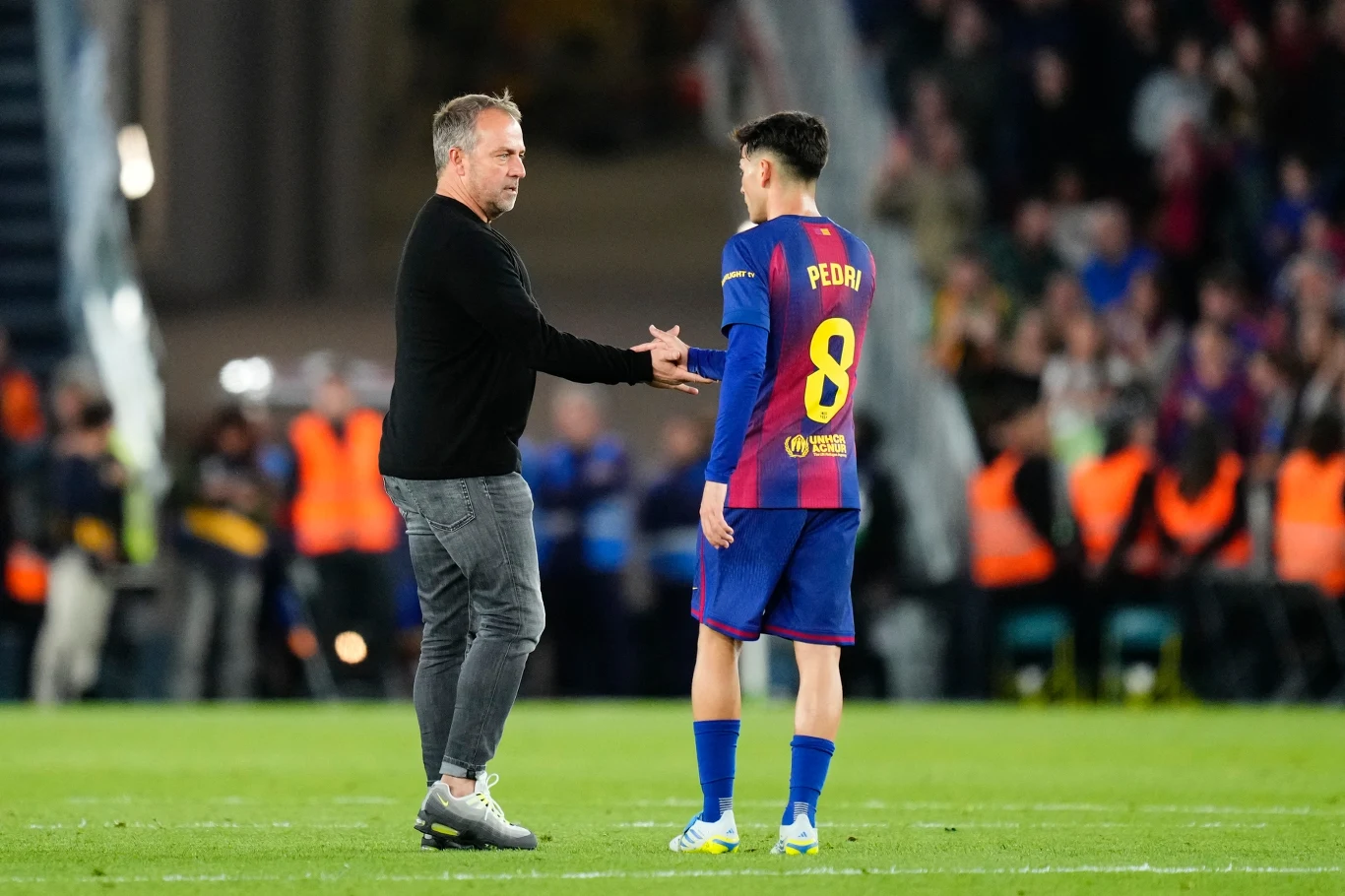 trener ubrany w czarny sweter i jeansy podaje rękę młodemu piłkarzowi w stroju FC Barcelony z numerem 8 i nazwiskiem Pedri, na stadionie z widoczną grupą stewardów w odblaskowych kamizelkach w tle