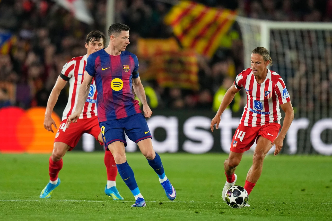 piłkarz FC Barcelony w granatowo-bordowej koszulce rywalizuje o piłkę z zawodnikiem Atletico Madryt w pasiastym czerwono-białym stroju podczas meczu na stadionie, w tle rozmyte flagi kibiców i trybuny pełne widzów