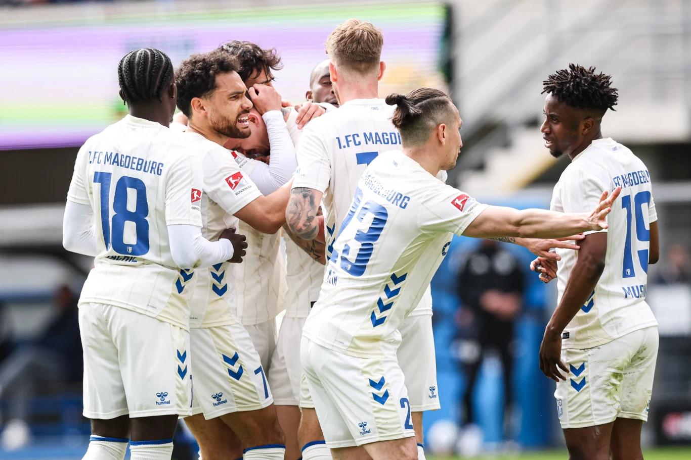 grupa piłkarzy w białych strojach z napisem 1. FC Magdeburg świętuje razem zdobycie gola na boisku, piłkarze są uśmiechnięci i przytulają się, na pierwszym planie widoczne emocje i gesty radości