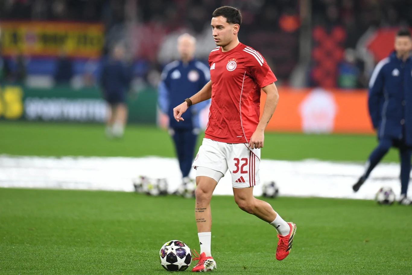 Santiago Hezze piłkarz w czerwonej koszulce i białych spodenkach z numerem 32 prowadzi piłkę nożną podczas rozgrzewki na stadionie, wokół niego kilka innych osób ubranych w stroje sportowe