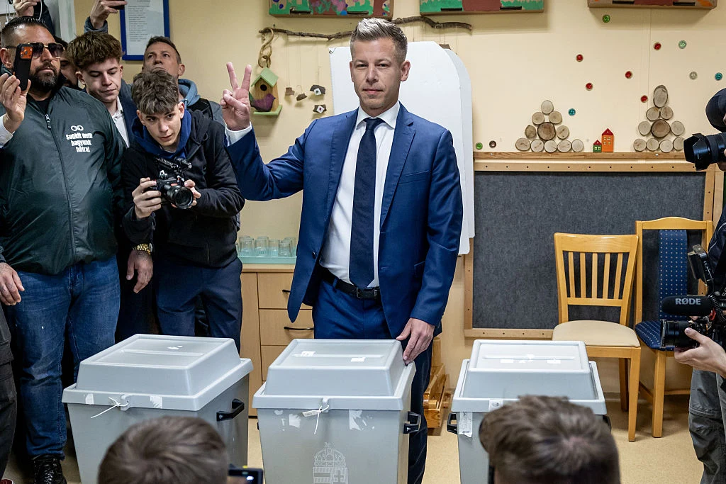 Peter Magyar podczas niedzielnego głosowania mężczyzna w garniturze stoi przy urnach wyborczych, unosi rękę z dwoma palcami w geście V, wokół niego zgromadzeni są fotoreporterzy i obserwatorzy