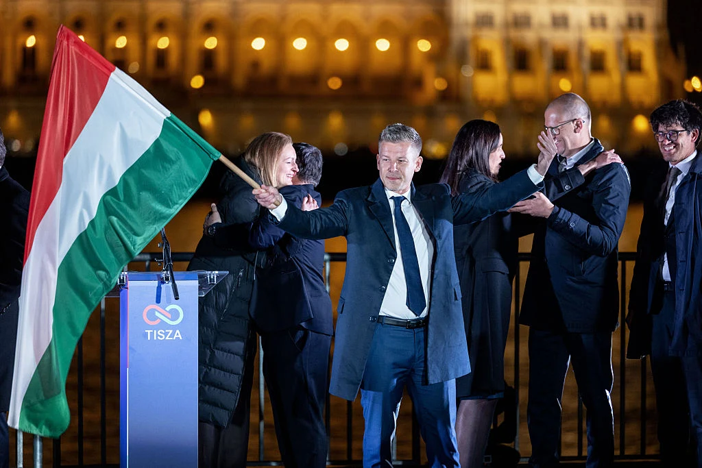 Mężczyzna w garniturze trzymający dużą węgierską flagę obok mównicy z napisem TISZA, otoczony przez grupę elegancko ubranych osób, które biją brawo i okazują wsparcie, w tle oświetlony budynek.