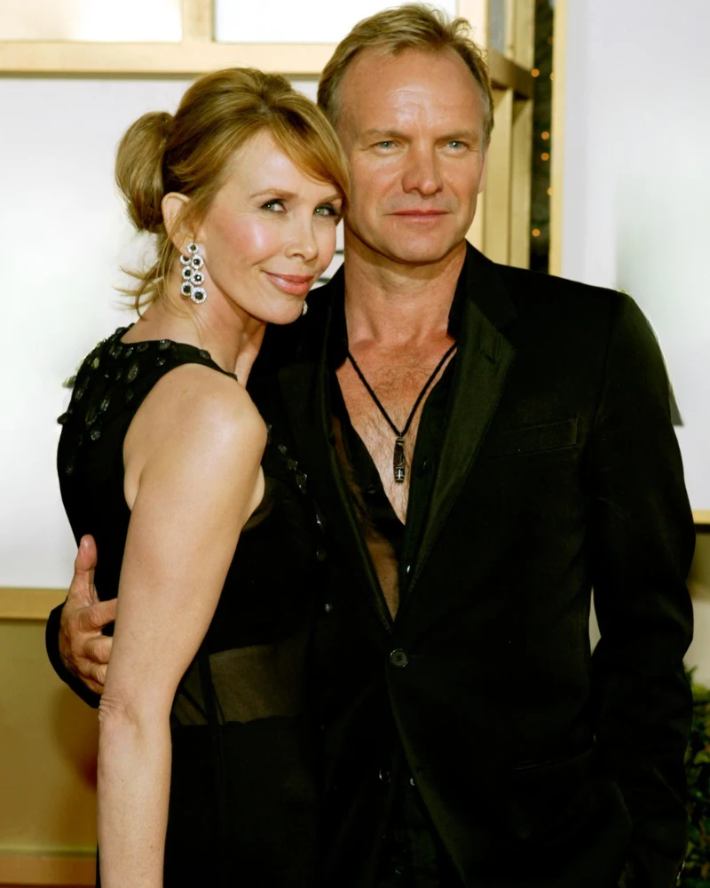 Para elegancko ubranych osób stoi blisko siebie, kobieta ma na sobie czarną sukienkę i efektowne kolczyki, mężczyzna ubrany jest w czarny garnitur z rozpiętą koszulą i zawieszką na szyi, oboje patrzą w stronę aparatu, prezentując się z klasą i pewności...