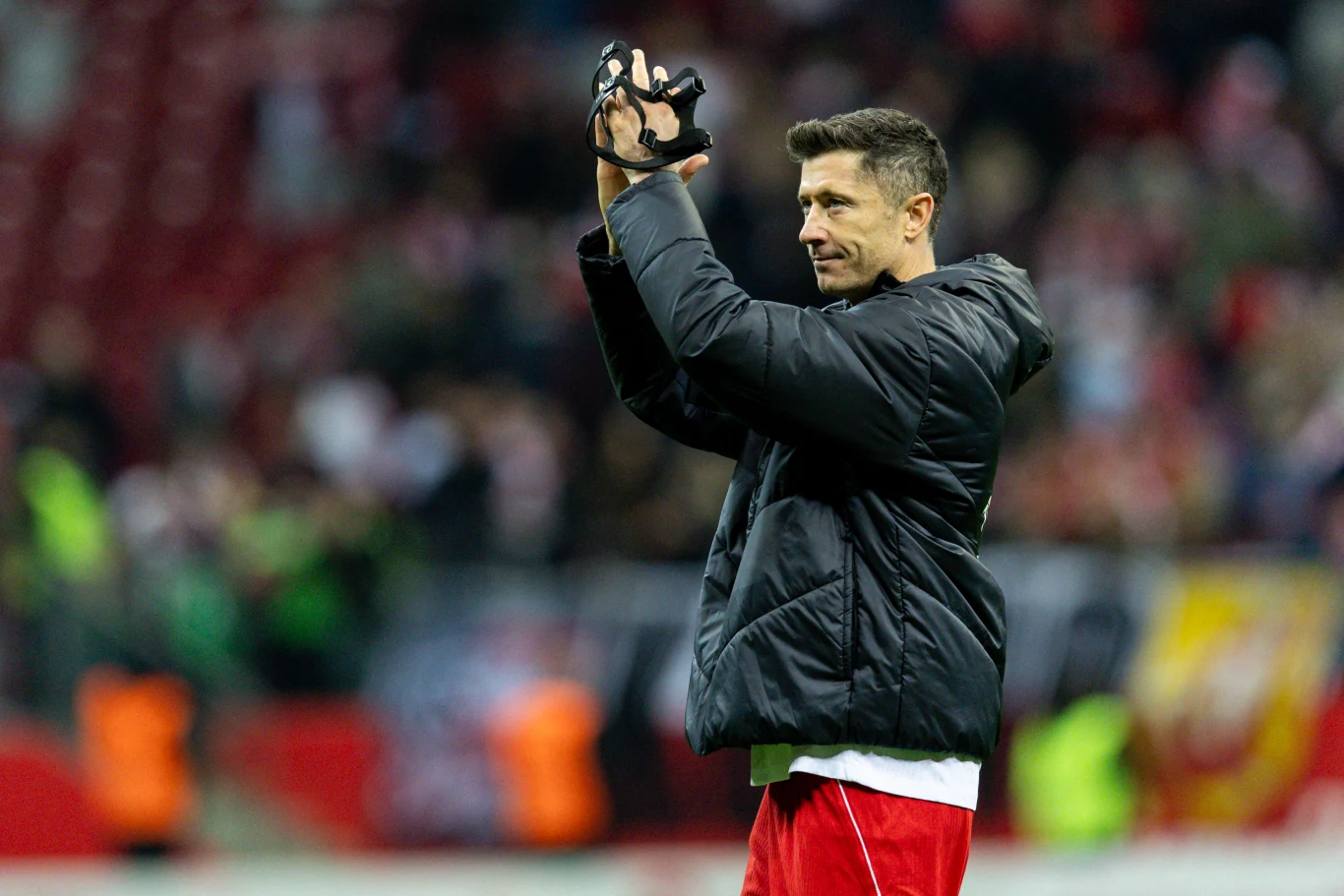 Robert Lewandowski Piłkarz w kurtce sportowej i czerwonych spodenkach klaszcze nad głową, trzymając w dłoniach czarną opaskę, na tle rozmytej publiczności na stadionie.