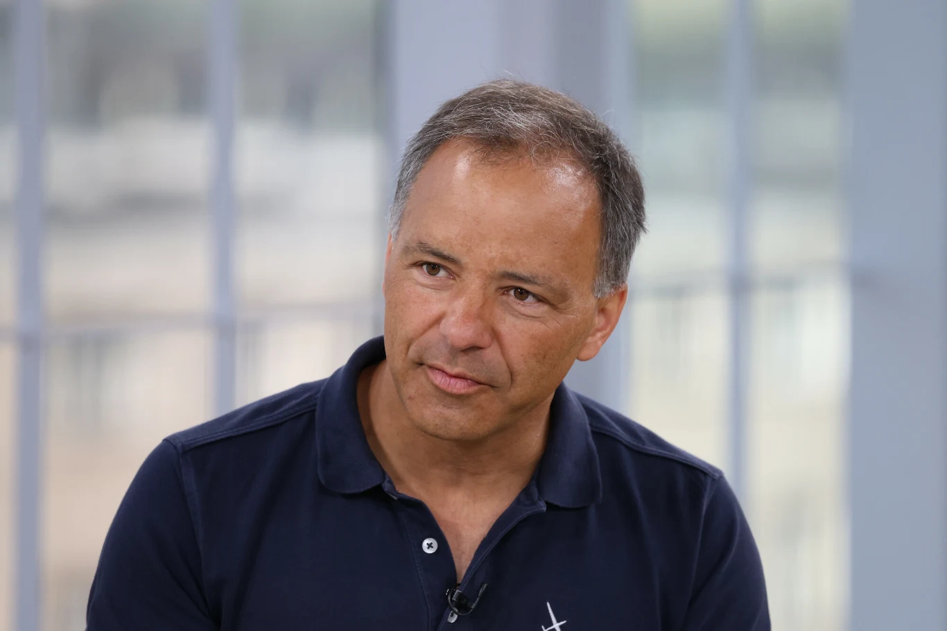 Sebastian Kawa Mężczyzna w średnim wieku o siwych włosach, ubrany w granatową koszulkę polo z przypiętym mikrofonem, siedzi na tle rozmytego, jasnego okna.