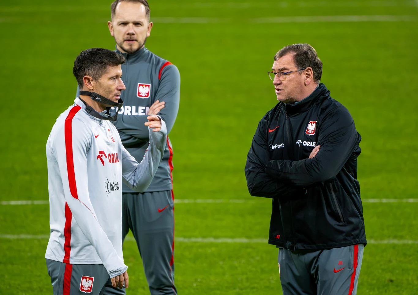 Robert Lewandowski i Jan Urban Trzech mężczyzn w sportowych dresach z emblematem polskiej reprezentacji piłkarskiej na tle zielonego boiska, dwóch z nich rozmawia ze sobą, a trzeci obserwuje sytuację z tyłu.