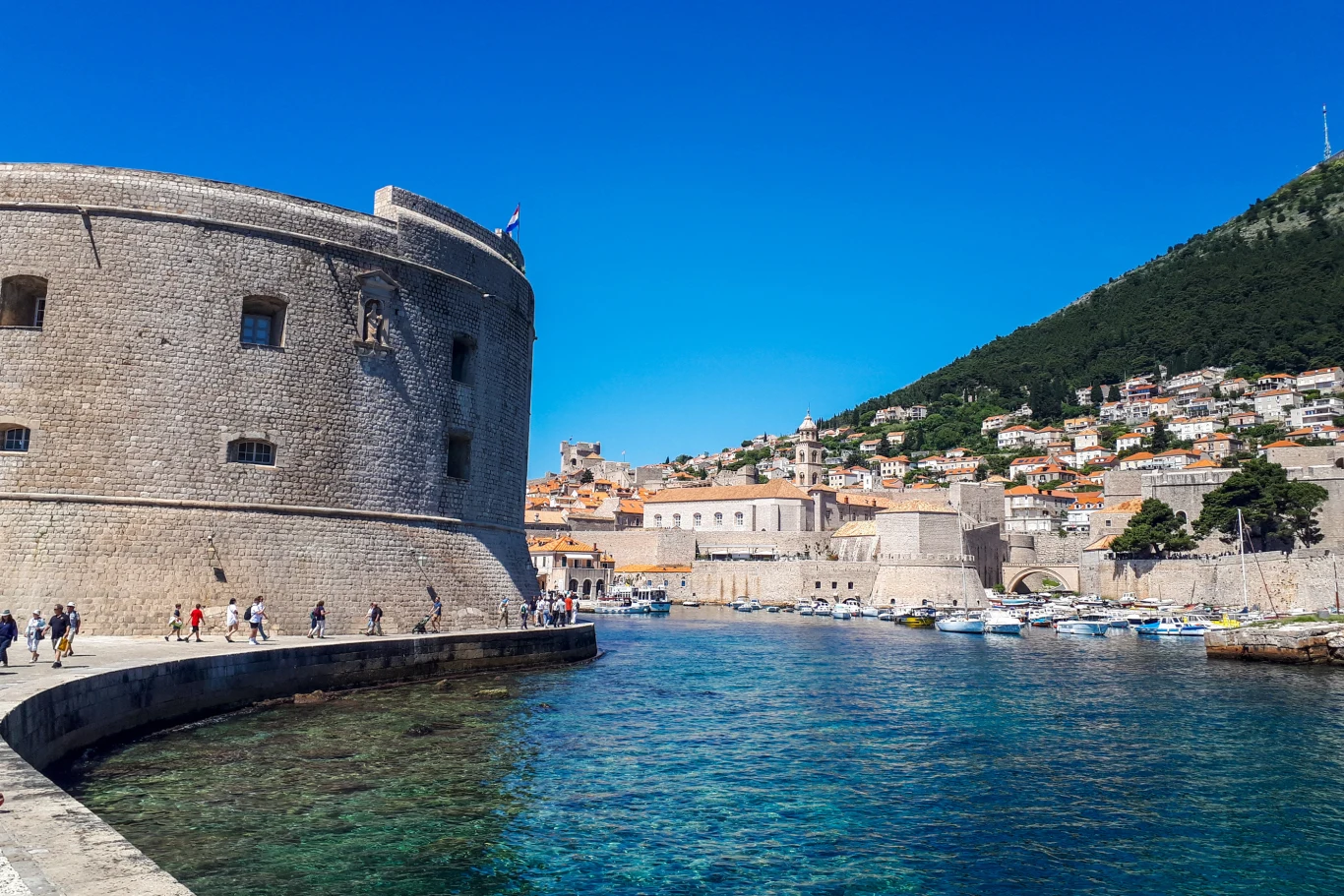 masywna kamienna twierdza przy brzegu morza, z wieloma turystami spacerującymi wzdłuż muru; po prawej stronie liczne białe łodzie zacumowane w porcie, w tle wzgórze zabudowane gęsto niskimi, jasnymi budynkami oraz intensywnie błękitne niebo
