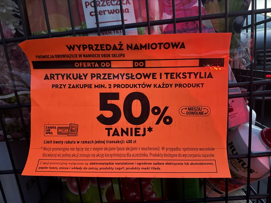 pomarańczowa informacyjna tabliczka z napisem o wyprzedaży namiotowej i rabacie 50% na artykuły przemysłowe i tekstylia przy zakupie minimum dwóch produktów, z wyróżnioną dużą liczbą procentową oraz warunkami promocji, umieszczona na metalowych kratkach