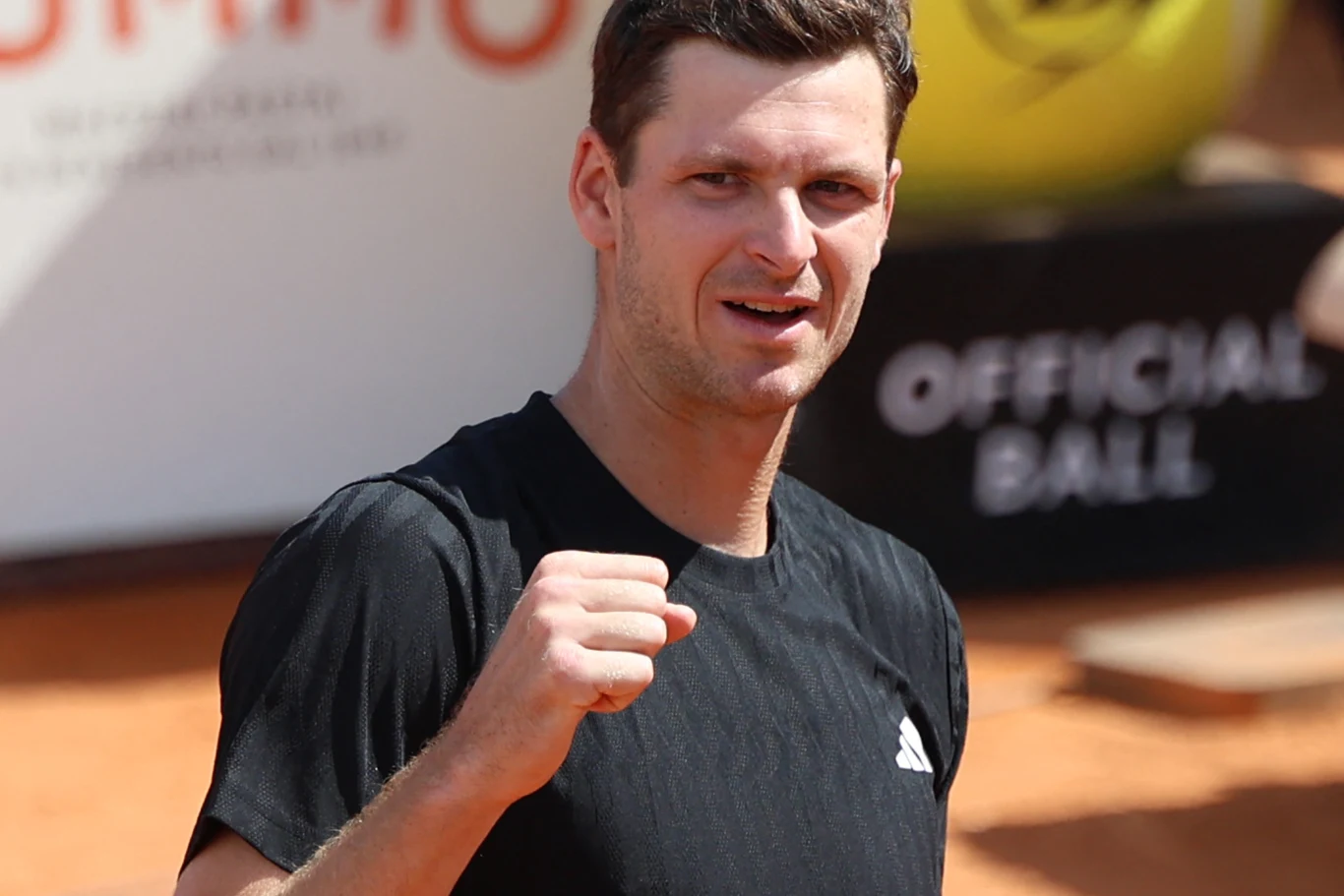 Mężczyzna w czarnej koszulce sportowej unosi zaciśniętą pięść w geście triumfu, w tle widoczne fragmenty kortu tenisowego i duża żółta piłka z napisem.