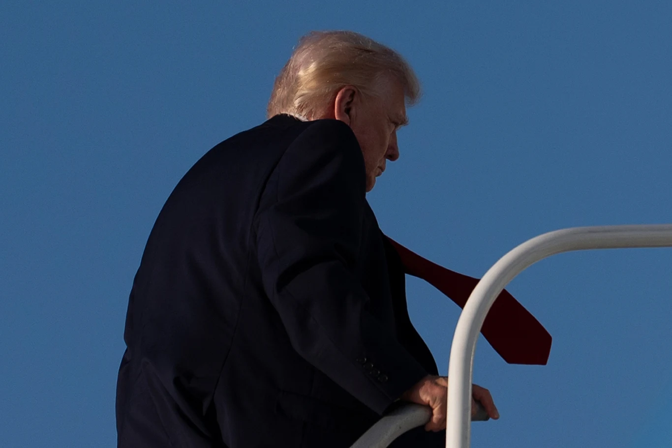 Donald Trump zmaga się z kryzysem - militarnym, energetycznym, wizerunkowym Mężczyzna w garniturze z czerwonym krawatem wchodzi po schodach na tle błękitnego nieba, z uchwyconym profilem twarzy i blond włosami podświetlonymi światłem.