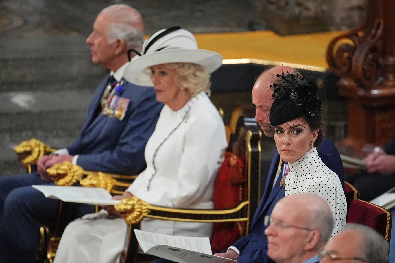 Cztery osoby w eleganckich strojach siedzą w rzędzie ozdobionych złotem foteli; kobieta w białym kapeluszu i mężczyzna w mundurze z medalami patrzą przed siebie, obok nich kobieta w biało-czarnej stylizacji z ozdobnym fascynatorem kieruje wzrok w stron...