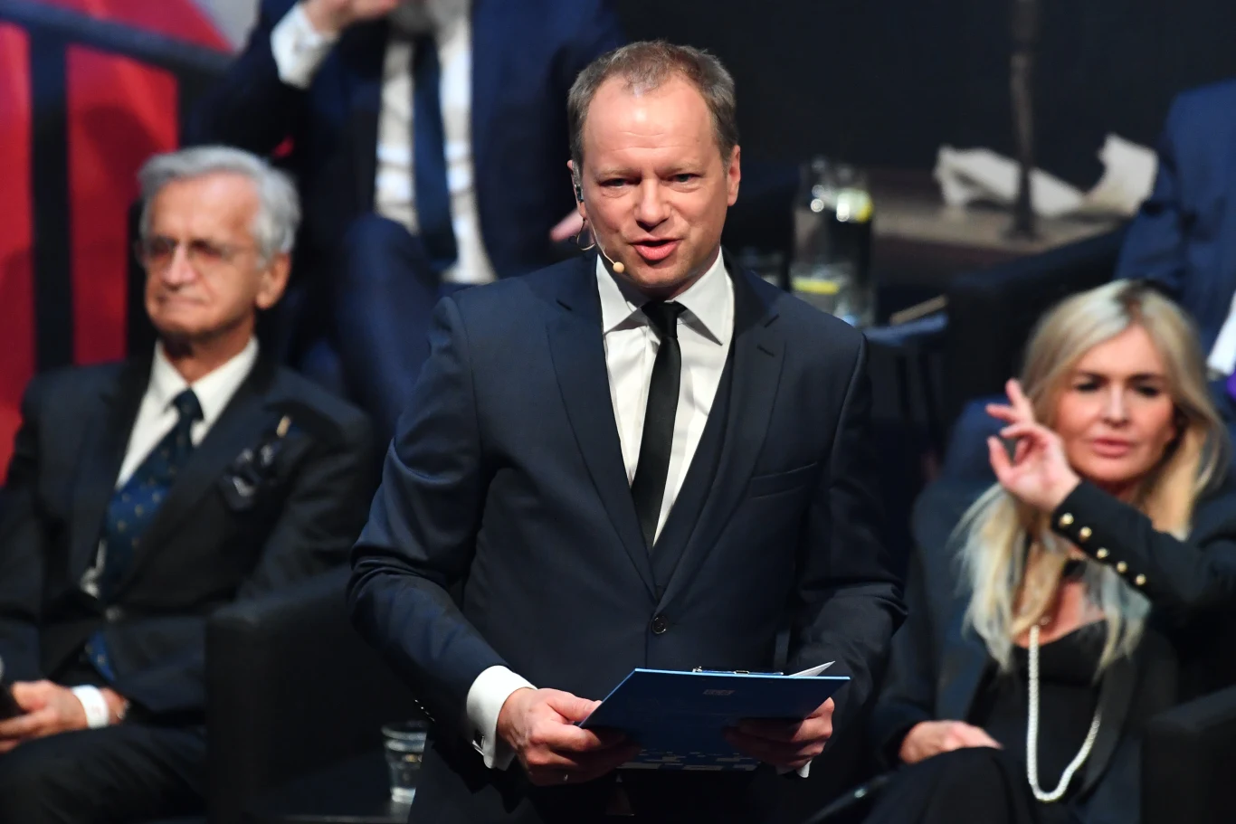 mężczyzna w garniturze z mikrofonem przypiętym do twarzy trzyma dokumenty, siedzący z tyłu starszy mężczyzna w marynarce oraz kobieta w jasnych włosach wykonująca gest ręką, wszyscy w eleganckich strojach, atmosfera oficjalnego wydarzenia lub gali