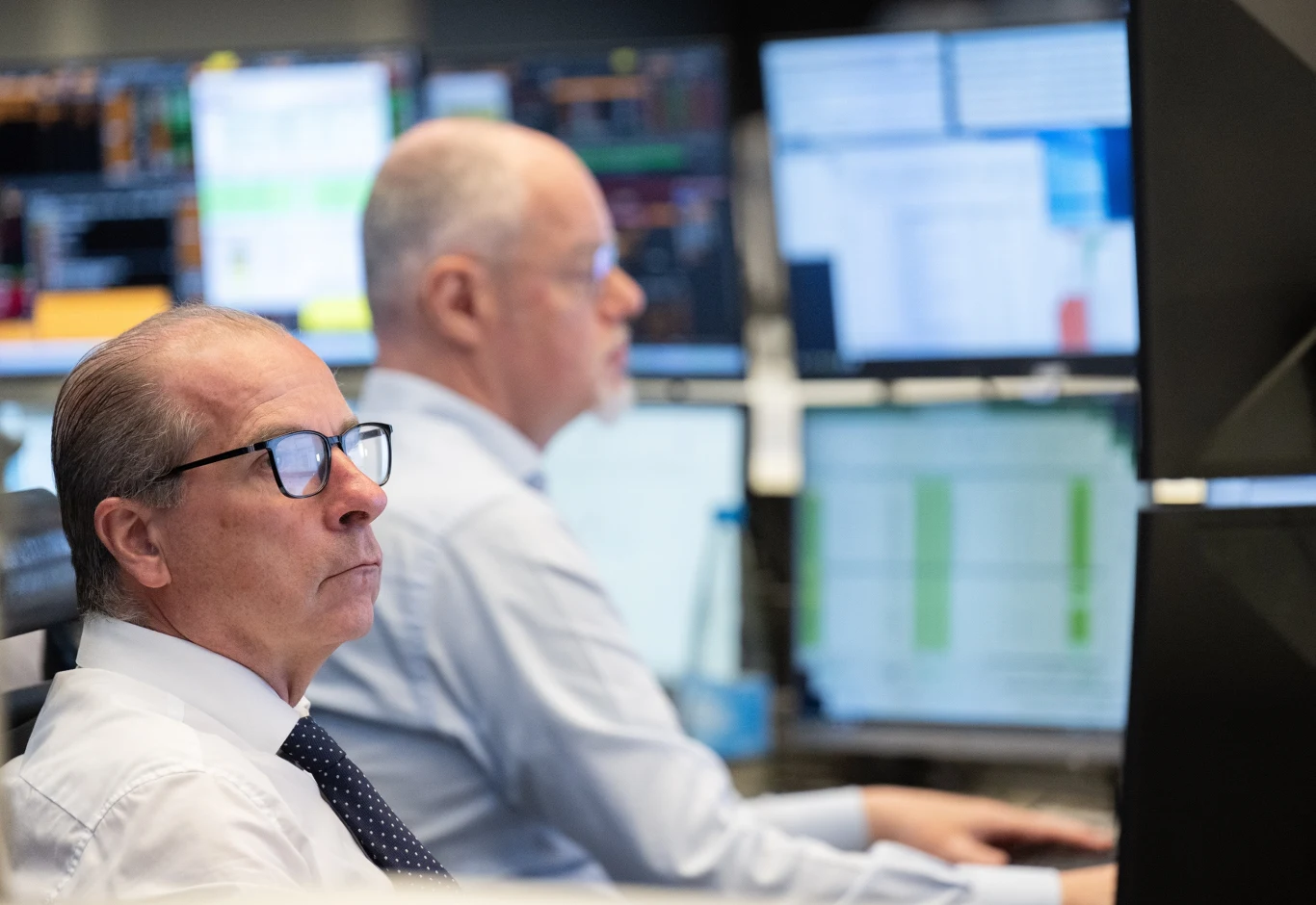 dwóch mężczyzn w koszulach siedzi przy stanowiskach komputerowych z wieloma monitorami, skoncentrowani na analizie wykresów i danych wyświetlanych na ekranach