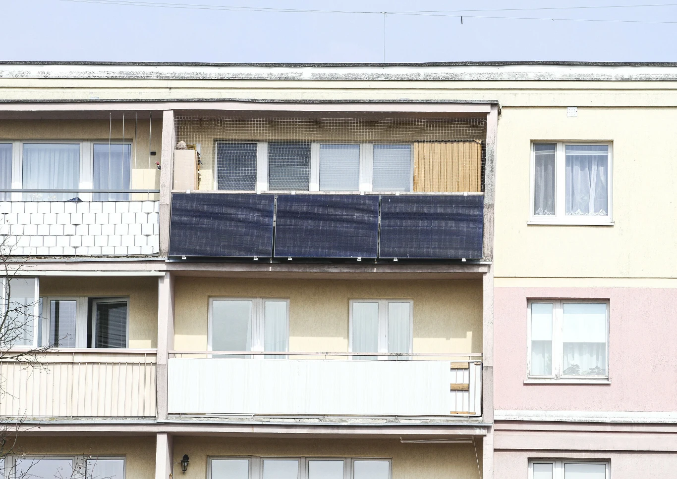 Kto może otrzymać dofinansowanie na na instalacje fotowoltaiczne i magazyny energii? Balkon w bloku z trzema panelami fotowoltaicznymi na balustradzie, jasna elewacja, siatka ochronna