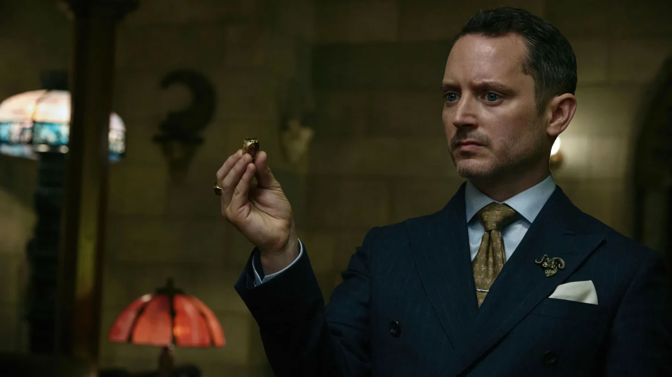 Elijah Wood w filmie "Zabawa w pochowanego 2" mężczyzna w eleganckim granatowym garniturze i krawacie trzyma w dłoni mały przedmiot, patrząc na niego z powagą; w tle lampy ze szklanymi abażurami i ściana z numerem 2