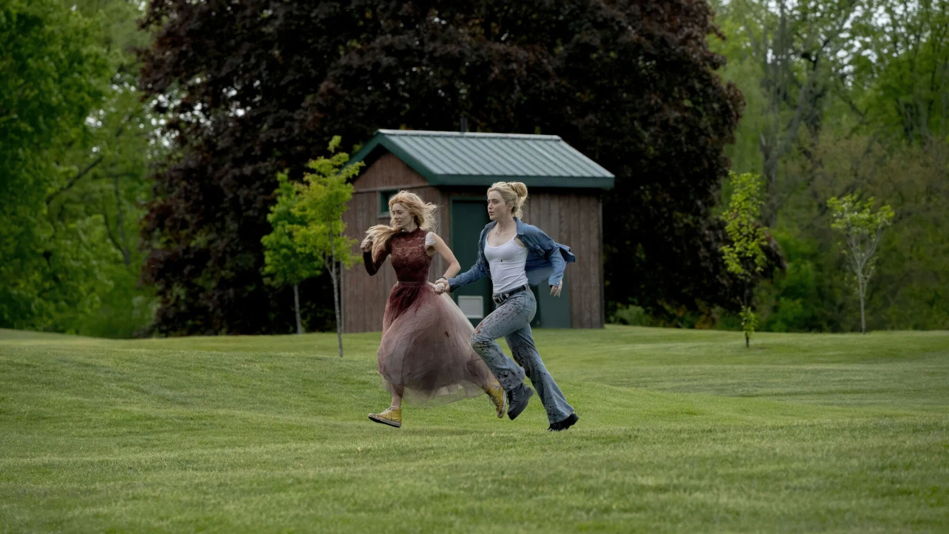 Samara Weaving i Kathryn Newton w filmie "Zabawa w pochowanego 2" dwie kobiety biegną przez zielony trawnik, jedna w bordowej sukni, druga w jasnej koszuli i jeansach, w tle drewniany budynek i drzewa