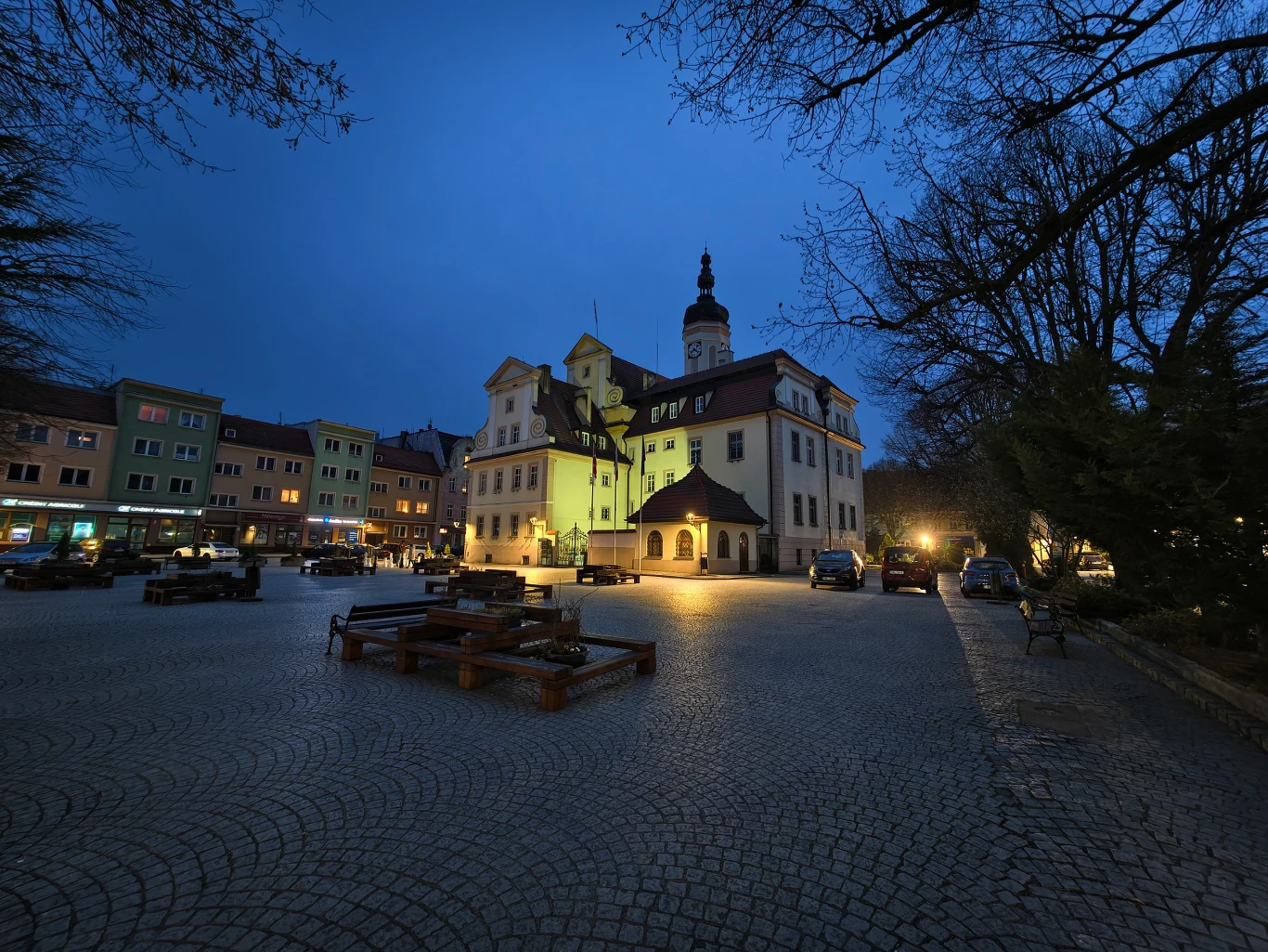 Zabytkowy ratusz oświetlony ciepłym światłem, otoczony brukowanym placem z ławkami i niskimi drzewami, w tle kamienice mieszkalne; wieczorna, spokojna atmosfera podkreślona granatowym niebem.