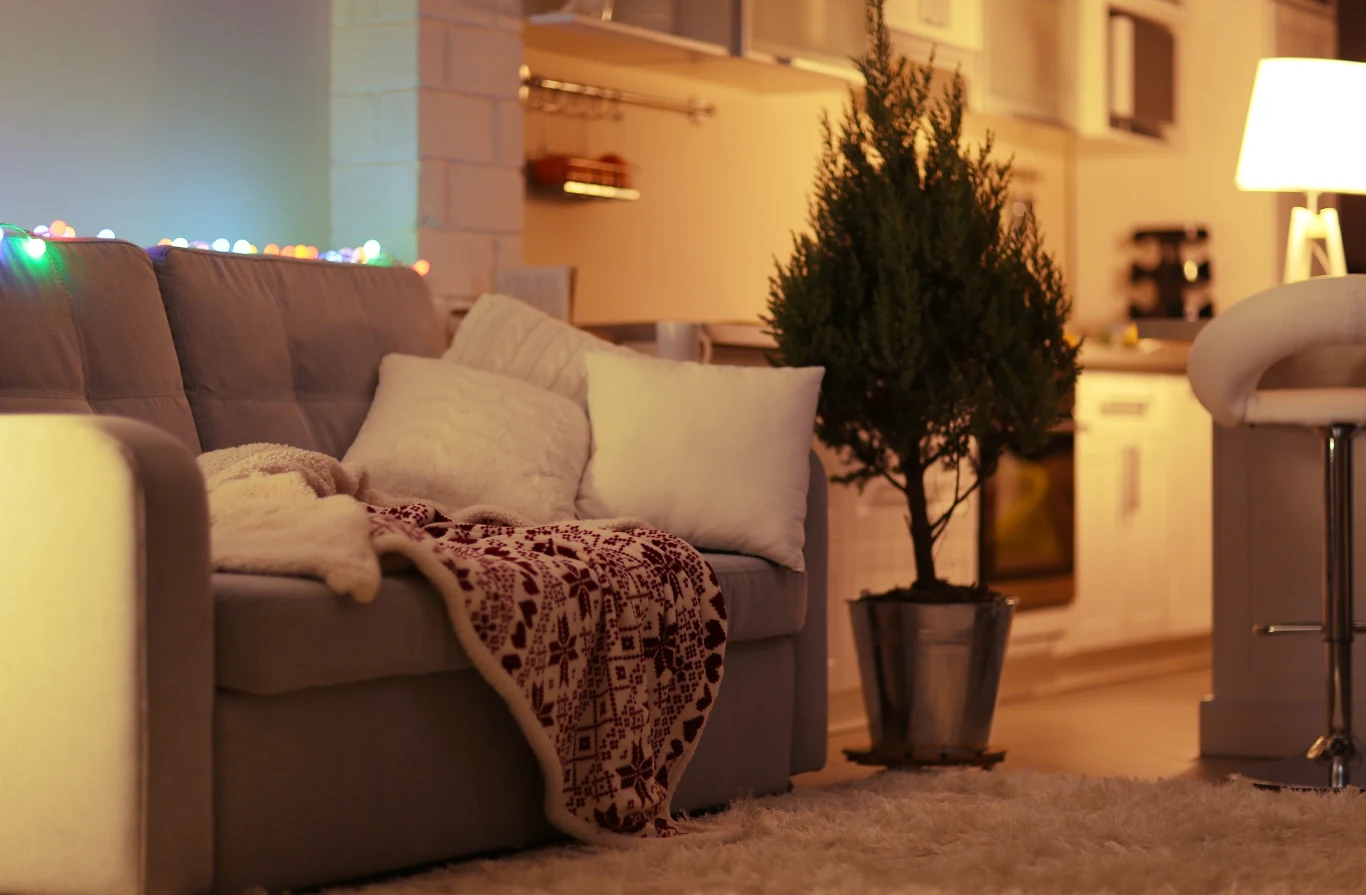 Szara sofa z białymi poduszkami i czerwonym kocem w świąteczny wzór, niewielka choinka w doniczce obok oraz kolorowe lampki na ścianie w przytulnym, nowoczesnym salonie połączonym z kuchnią.