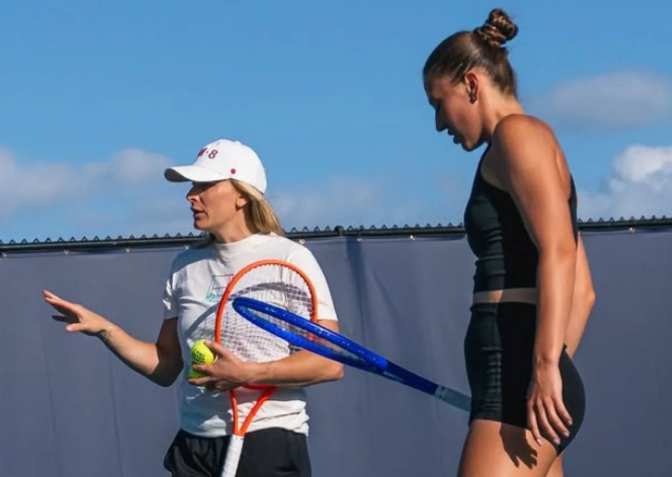 Sandra Zaniewska i Marta Kostiuk Trenerka w białej czapce oraz koszulce tłumaczy coś tenisistce ubrane w sportowy, czarny strój; obie stoją na korcie tenisowym na tle niebieskiego nieba, trenerka trzyma rakietę i piłki tenisowe, a zawodniczka słucha uważnie.