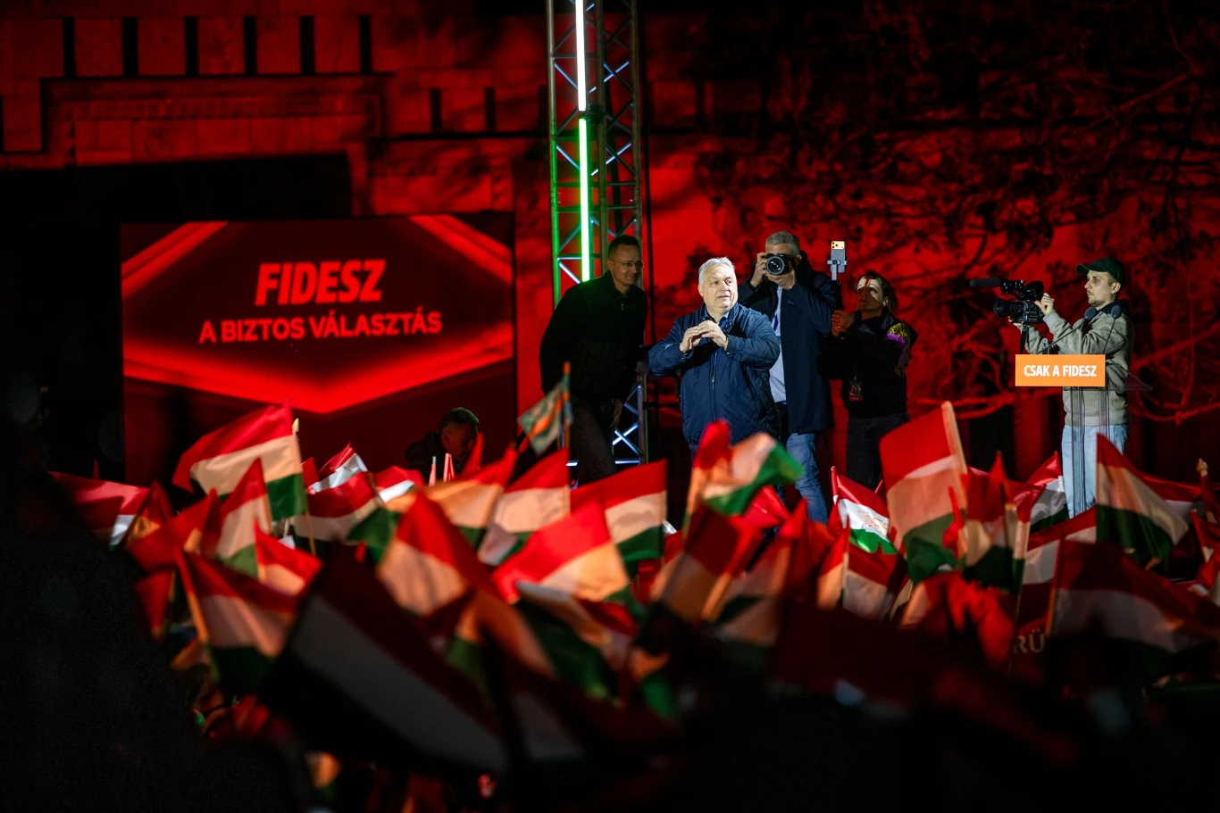 Viktor Orban tłum ludzi machających węgierskimi flagami przed sceną, na której przemawia mężczyzna, w tle podświetlona tablica z napisem 'FIDESZ A BIZTOS VALASZTÁS', scena i tłum oświetlone intensywnym czerwonym światłem, po prawej stronie osoby z kamerą i aparatem...