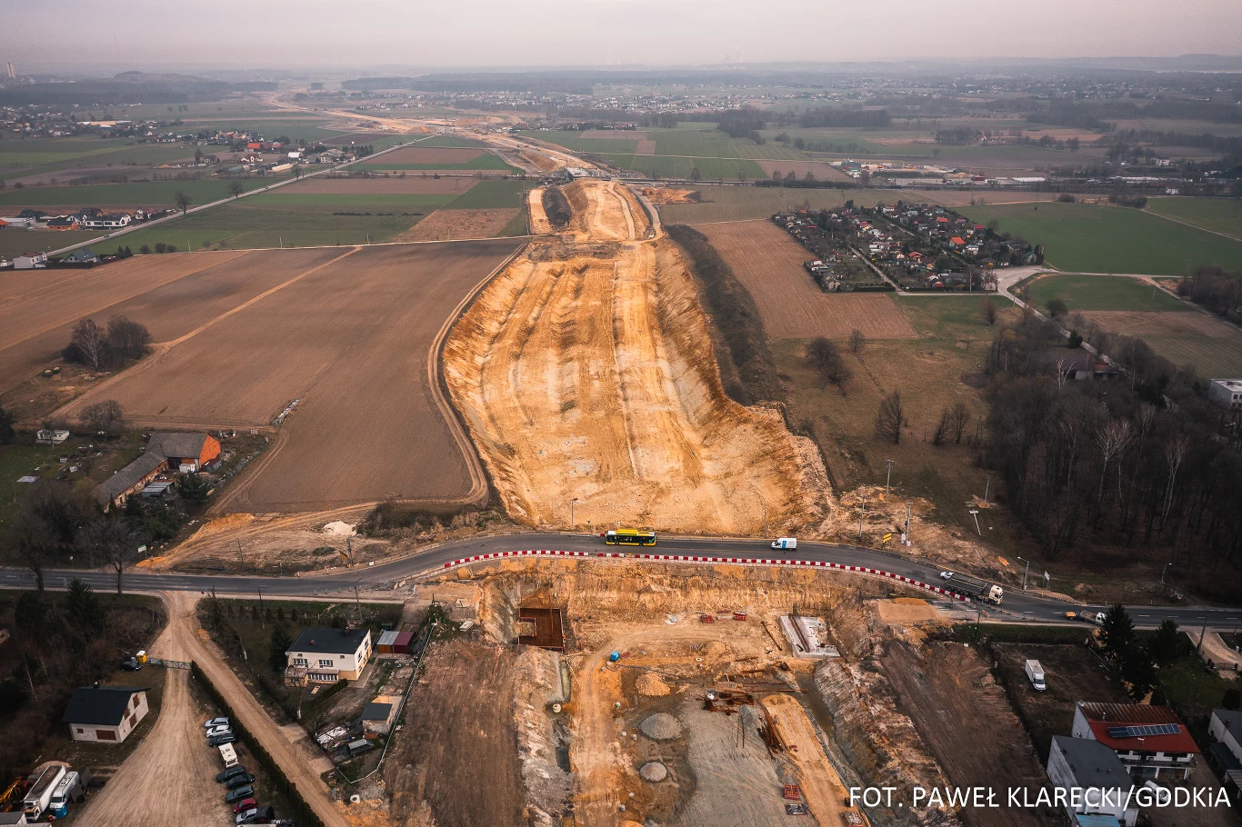Szeroki pas terenu przygotowany pod budowę drogi, otoczony polami uprawnymi oraz domami jednorodzinnymi, z widoczną przeciętą drogą lokalną i maszynami budowlanymi pracującymi przy inwestycji.