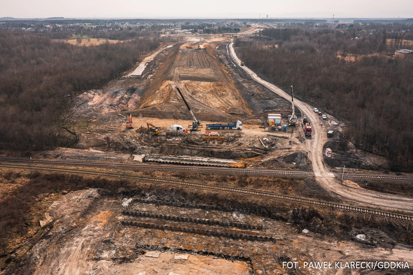 rozległy plac budowy nowej drogi przecinający leśny teren, na pierwszym planie tory kolejowe oraz maszyny budowlane i baraki na zapleczu technicznym, ziemia rozkopana i przygotowywana pod infrastrukturę drogową