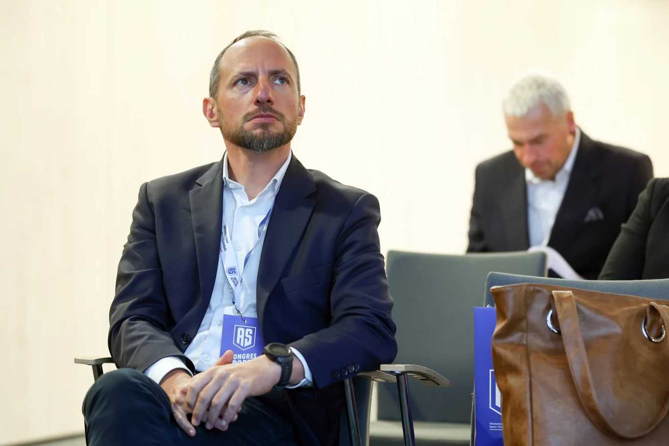Mężczyzna w garniturze siedzi na krześle z identyfikatorem na szyi, obok niego na krześle znajduje się brązowa torba, w tle widoczny starszy mężczyzna zajęty czytaniem dokumentów.