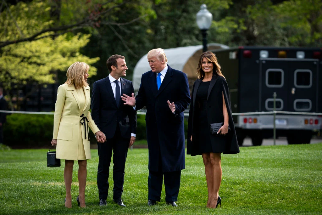 Podczas spotkania par prezydenckich USA i Francji w 2018 r., zarówno Melania Trump, jak i Brigitte Macron nosiły krótkie spódnice Cztery elegancko ubrane osoby stoją na zielonym trawniku, rozmawiając i uśmiechając się do siebie; dwie kobiety w jasnym płaszczu i czarnej sukience oraz dwaj mężczyźni w garniturach i płaszczu tworzą centralną grupę na tle zielonych drzew i zaparkowan...