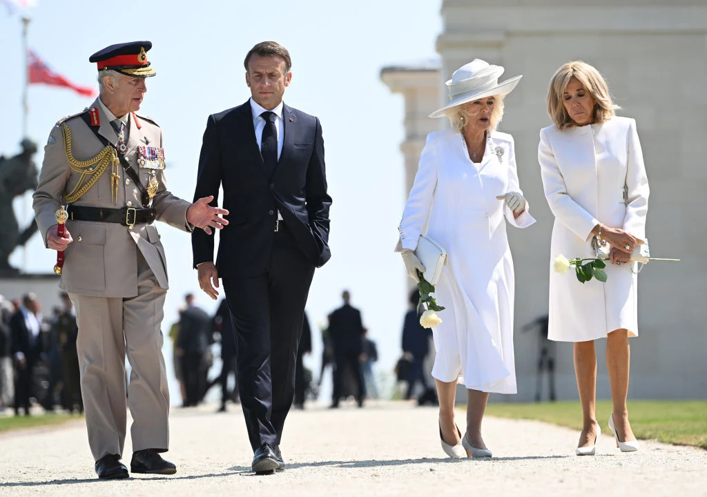 Brigitte Macron (71 l.) i królowa Kamila (77 l.) podczas spotkania w 2024 r. Cztery elegancko ubrane osoby idą razem aleją, dwie kobiety w białych strojach trzymają w dłoniach białe róże, mężczyzna w ciemnym garniturze oraz drugi w pełnym mundurze wojskowym prowadzą rozmowę, w tle jasny budynek oraz postacie spacerujące na słońcu.
