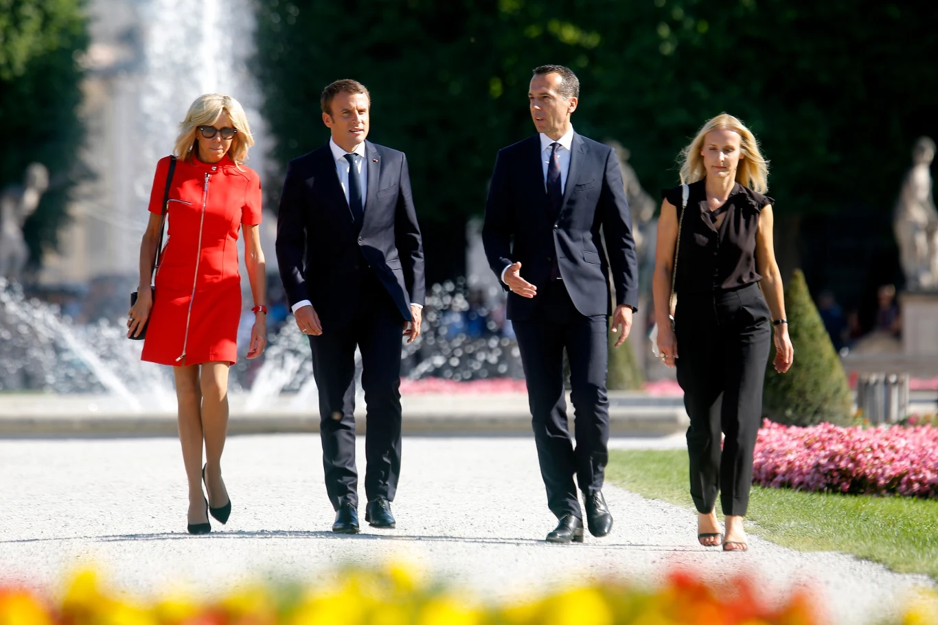 Brigitte Macron lubi czerwoną sukienkę od Louis Vuitton. Kilkukrotnie była w niej widziana. Tutaj podczas wizyty w Austrii w 2017 r. Cztery elegancko ubrane osoby spacerują alejką ogrodową wśród kwiatów, w tle widoczne są fontanny i rzeźby ogrodowe.